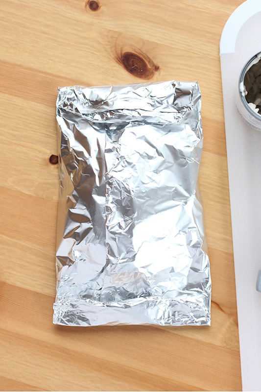 Top down image showing a closed tin foil dinner packet on a wooden table