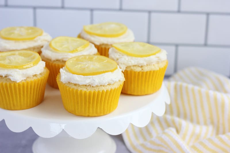 horizontal image of a white cupcake tray that has scalloped edges holding six baked and frosted coconut lemon cupcakes topped with candied lemon slices