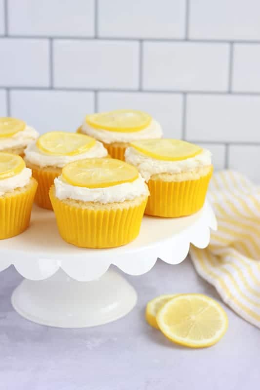 white cake stand with scalloped edges topped with lemon cupcakes in yellow liners with coconut frosting and a candied lemon slice on top of each