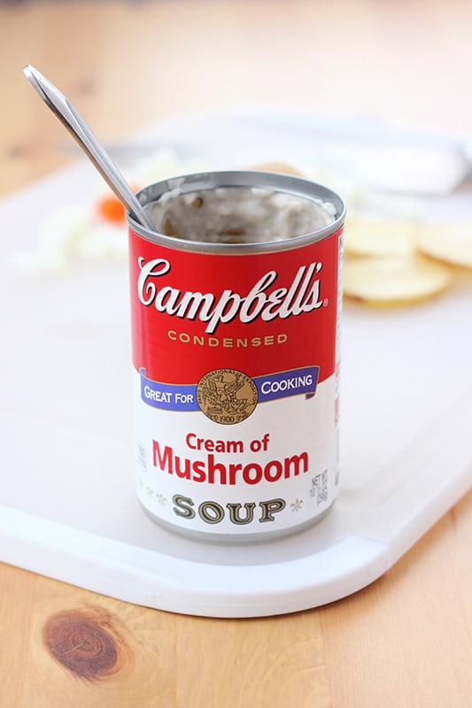 Can of cream of mushroom soup open with a spoon inside sitting on a white cutting board with sliced potatoes in the background
