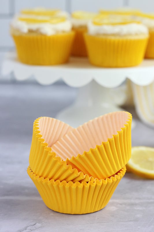 stack of yellow cupcake liners on a gray table top with baked cupcakes on a white scalloped edge cake stand in the background