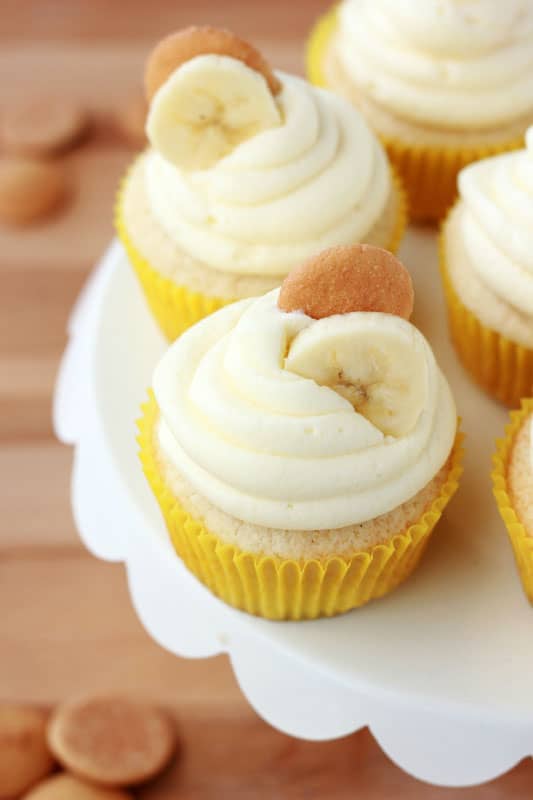 Close up image of a banana cupcake in a yellow wrapper topped with a swirl of banana pudding frosting and a vanilla wafer cookie with a banana slice