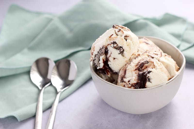 horizontal image of a bowl of ice cream with a chocolate fudge swirl throughout the scoops. Two spoons and a light blue napkin laying off to the side. 