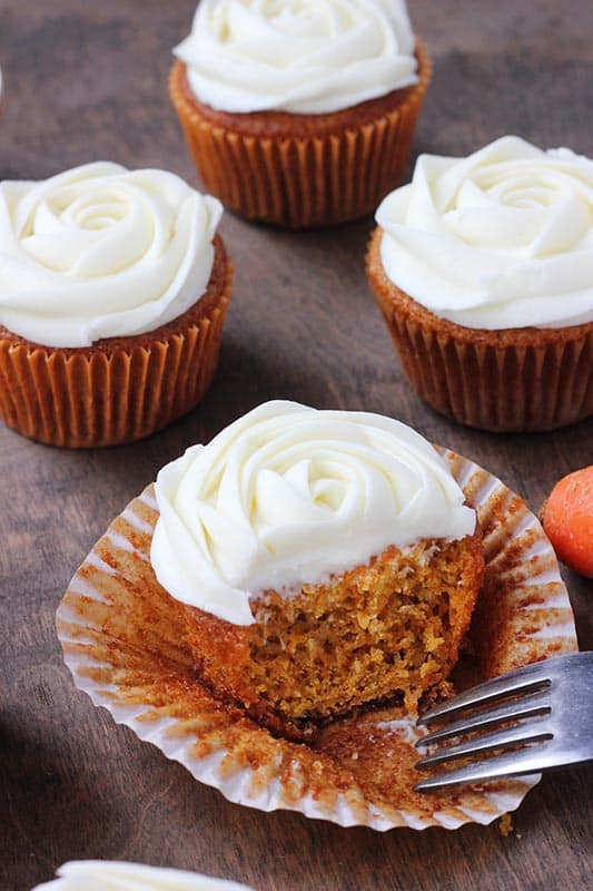 carrot cupcake that is sitting inside of an open wrapper with white cream cheese frosting piped on top to look like a flower with a bite sitting on a fork