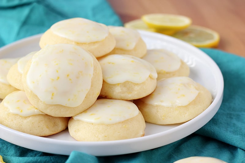 close up image showing a white plate full of lemon cookies with a lemon glaze sitting on top of a teal napkin with lemon slices off in the background