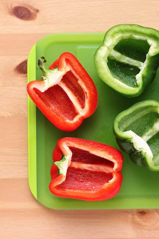 green cutting board sitting on a wooden table top with red and green peppers that have been sliced in half and cleaned of seeds