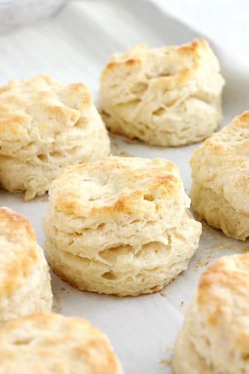 close up image of a baked biscuit from the side showing the fluffy layers. Biscuit is sitting on a baking tray that is covered in parchment paper