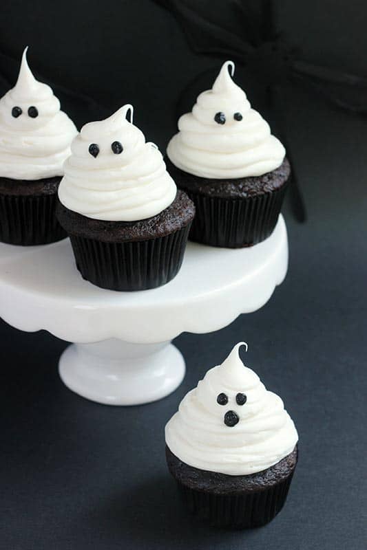 white cake stand with slightly scalloped edges sitting on a black backdrop and topped with three ghost decorated cupcakes with one cupcake on the bottom off to the side