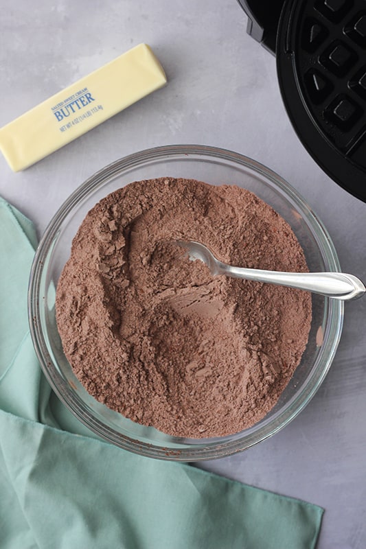 top down image of a glass mixing bowl with a chocolate powder sitting on a white table top with a light blue napkin and a stick of butter sitting off to the side