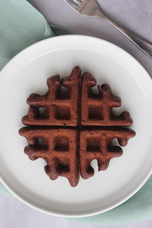 top down image of a dark brown waffle sitting on a white round plate with a lifted edge on top of a light blue napkin with a fork sitting off to the side