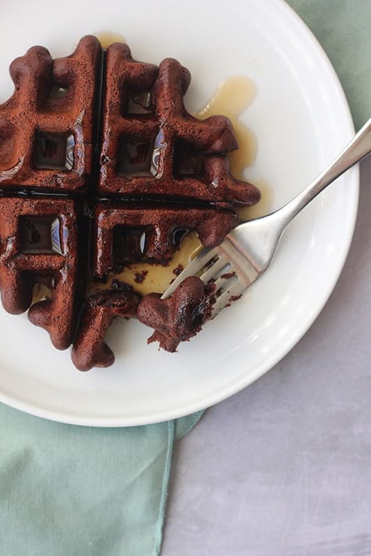 close up image of a chocolate waffle sitting on a white plate with syrup on top and a bite sitting on a fork. A light blue napkin is off to the side