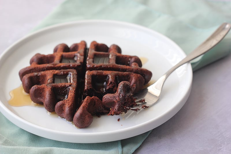 side view image of a dark brown waffle sitting on a white round plate with a lifted edge on top of a light blue napkin with a fork sitting off to the side