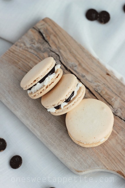 S'more macarons sitting on a wooden plank with chocolate chips sprinkled on the table next to the plank