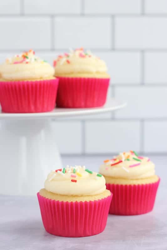 Two frosted vanilla cupcakes topped with sprinkles and wrapped in bright pink cupcake liners on a countertop, next to more cupcakes on a cake stand in the background.