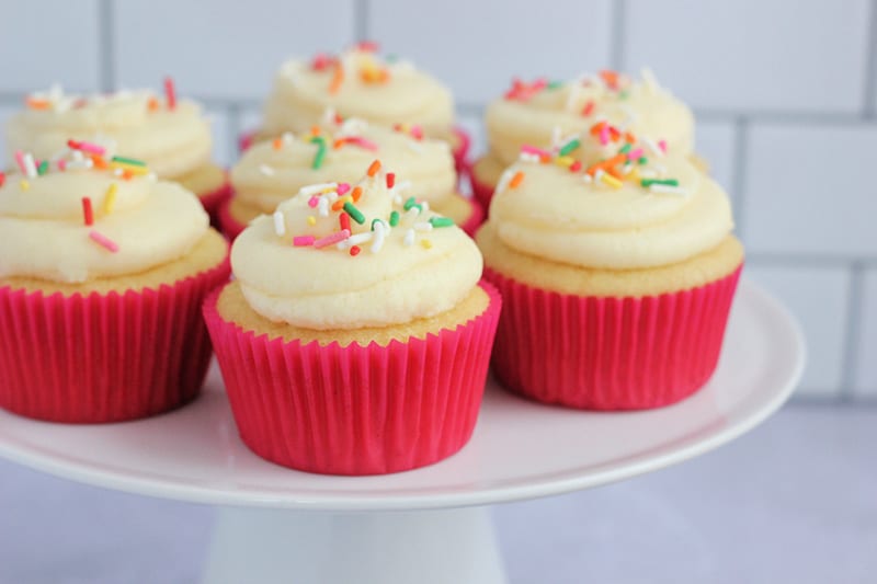 Frosted vanilla cupcakes topped with sprinkles, wrapped in bright pink cupcake liners on a cake stand.