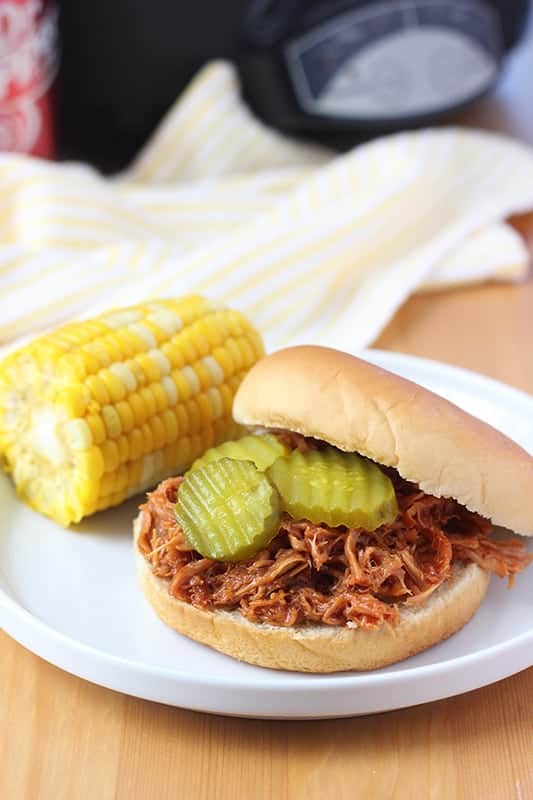 Pulled Pork Sandwich on white plate with pickles and side of corn on the cob