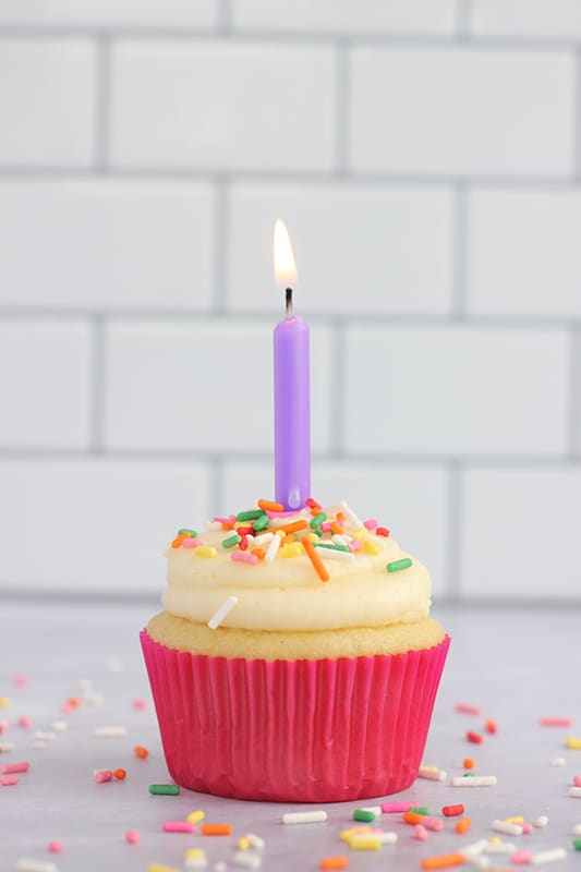 A frosted vanilla cupcake topped with sprinkles and a lit birthday candle, wrapped in a bright pink cupcake liner on a countertop.