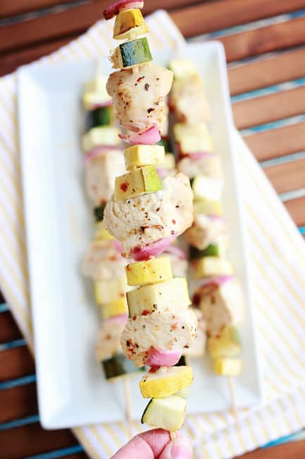 hand holding a skewer that is loaded with chicken, red onion, zucchini, and yellow squash over a plate of other skewers