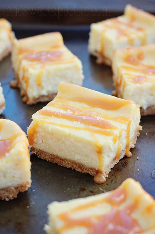 close up image showing a thick cheesecake bar that has been drizzled in a caramel sauce. Other bars are scattered around the first on a baking tin