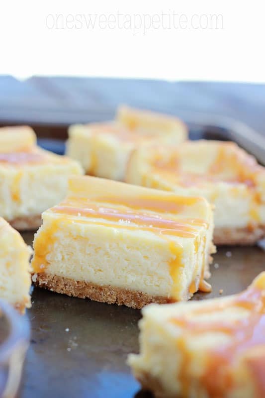 close up image showing a thick cheesecake bar that has been drizzled in a caramel sauce. Other bars are scattered around the first on a baking tin