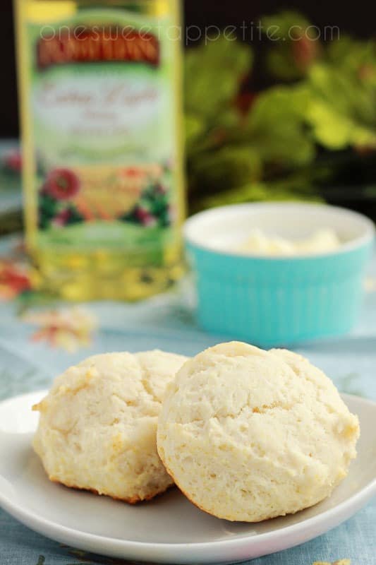 two biscuits sitting on a small white plate on a floral table cloth with butter and olive oil off in the background 