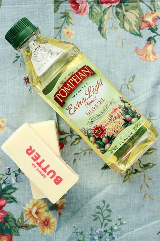 top down image showing a container of olive oil with two sticks of butter on a floral tablecloth 