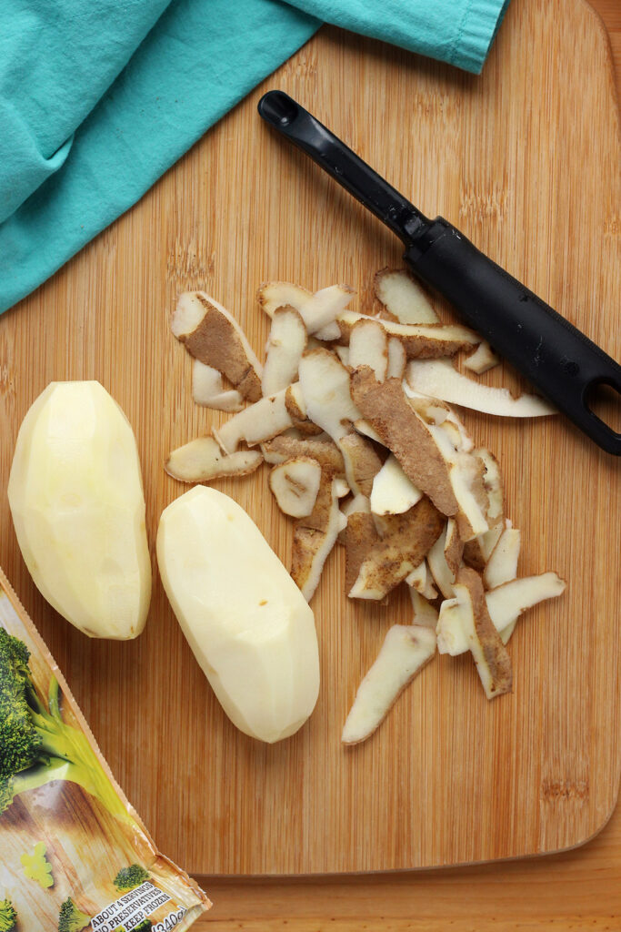 two peeled potatoes, with the peels on the side, with a potato peeler on a wooden cutting board with a teal napkin
