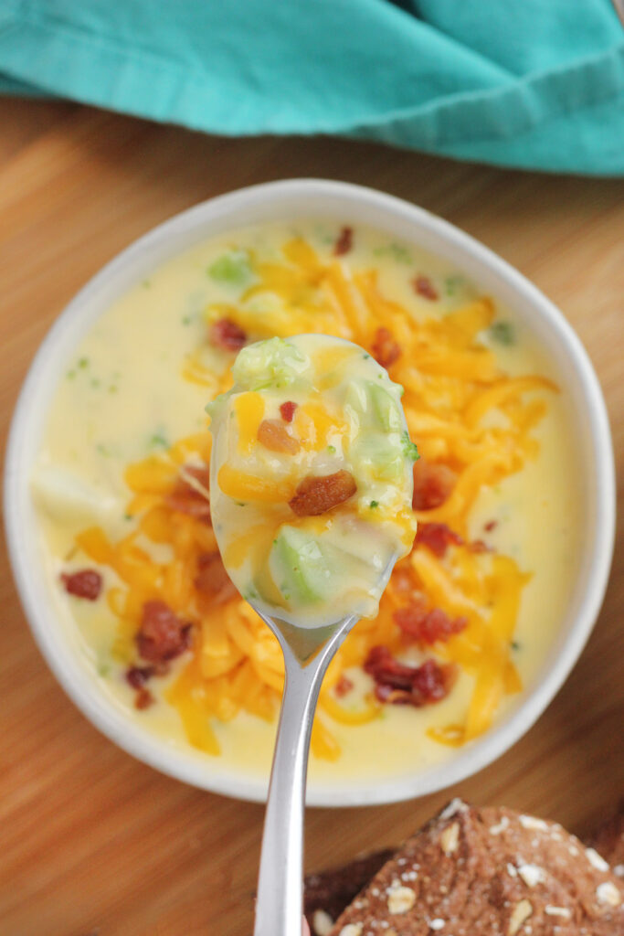 bowl of broccoli soup that is topped with shredded cheese and bacon pieces. A spoonful is being held above the bowl.