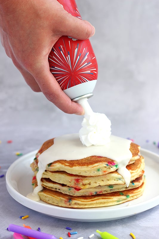 stack of funfetti pancakes on a white plate with frosting drizzling over the sides. Hand holding a whipped cream can on top spraying cream onto the frosting