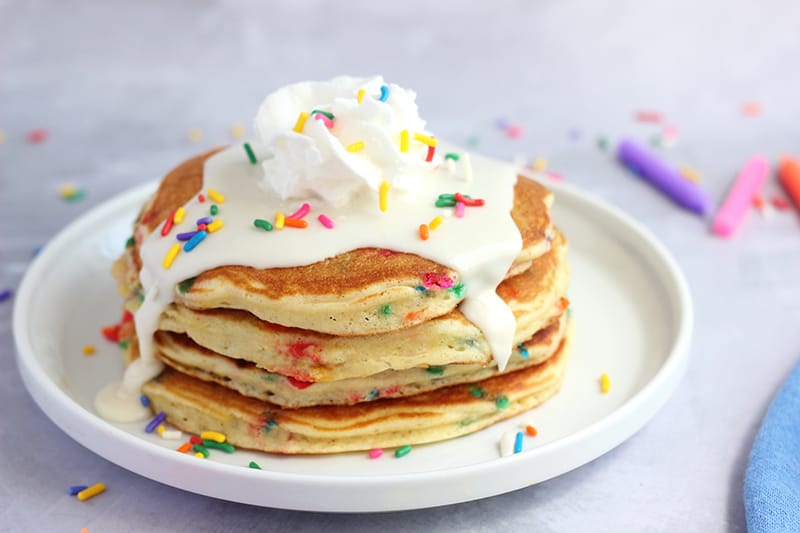 stack of four pancakes that are filled with sprinkles stacked on a white plate and topped with a drizzle of frosting, whipped cream, and more sprinkles