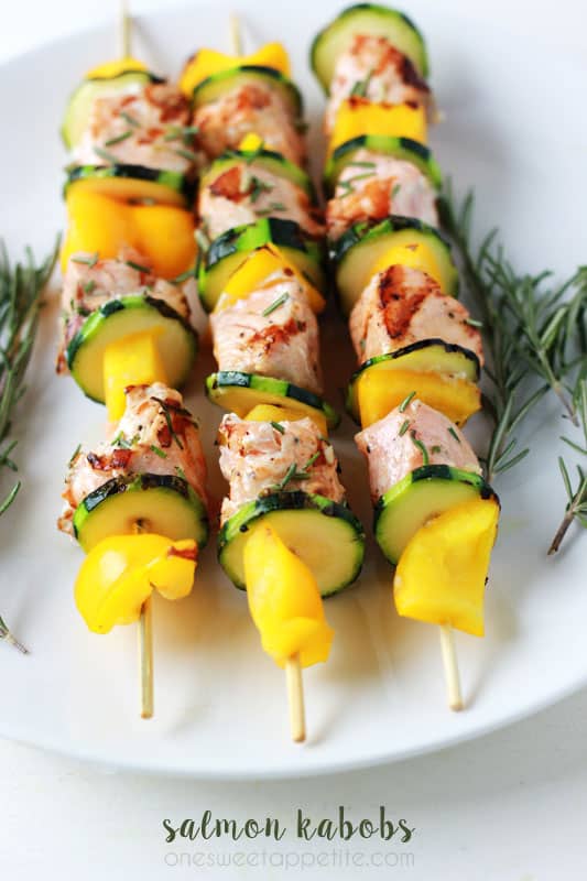 three salmon kabobs on a white plate with rosemary on the side