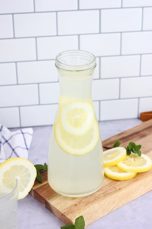 short textured glass filled with a lemon flavored drink with a lemon slice on the edge of the glass. A small sprig of mint is sitting off to the side of the glass with a wooden cutting board and pitcher of more drink. There are also lemon slices off in the background