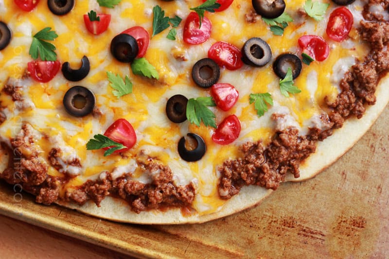 top down close up image of a pizza that is topped with ground beef, cheese, olives, tomatoes, and cilantro on a baking tray