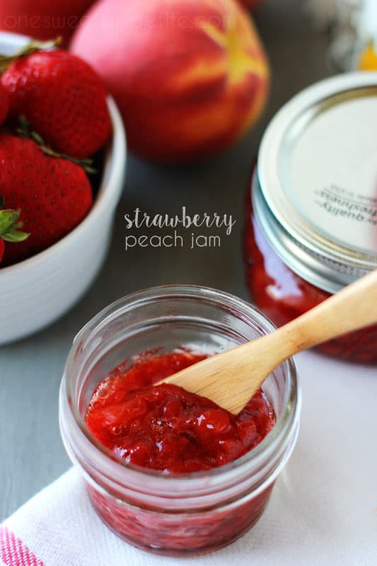 small jar of red jam with a wooden knife sitting inside the container. Jar is sitting on a white napkin on a gray table top. Fresh strawberries, peaches and other jars of jam sitting off to the side