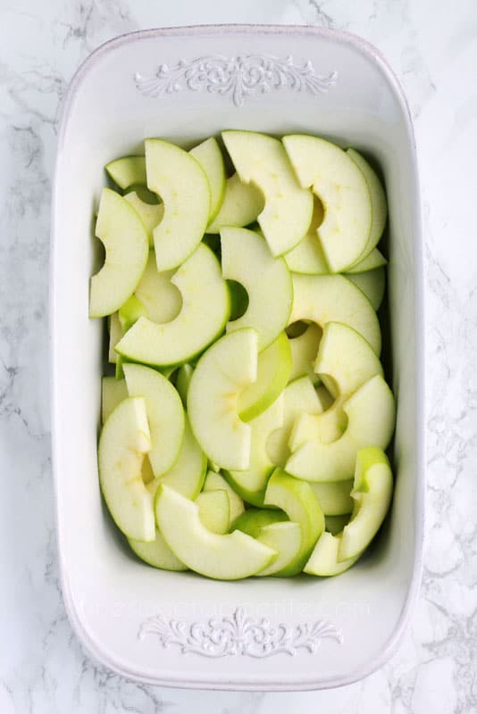 top down image showing sliced green apples sitting in a white baking pan on a white marble counter. 