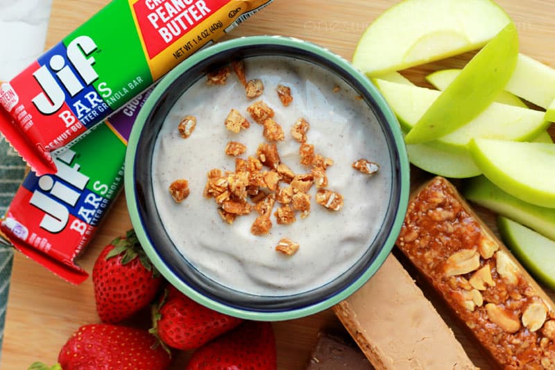 top down image showing a blue bowl filled with a cinnamon dip topped with granola crumbles. Around the bowl are sliced apples, granola bars, and fresh strawberries 