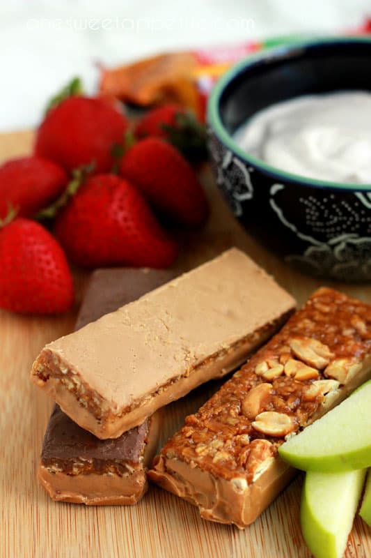 close up image showing a stack of peanut butter granola bars on a wooden cutting board with sliced apples and fresh strawberries off to the side with a blue floral bowl of dip
