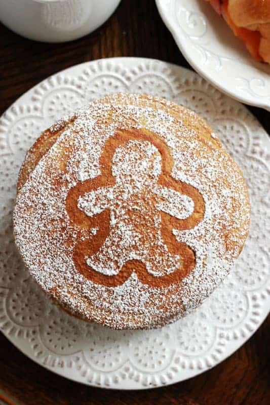 stack of gingerbread pancakes on a white plate with a dusting of powdered sugar in the shape of a gingerbread cookie