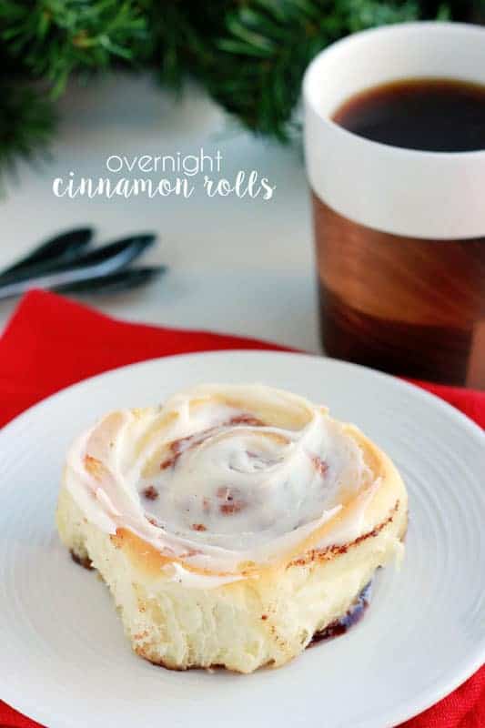 single cinnamon roll sitting on a white plate with a red napkin and a cup of hot chocolate to the side