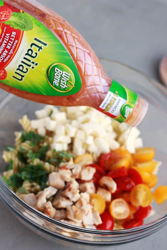 Italian dressing being drizzled over a bowl of fresh pasta ingredients on a grey table