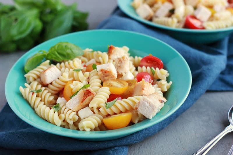 bright blue bowl filled with spiral pasta that is topped wtih grilled chicken, tomatoes, and fresh basil sitting on a blue napkin