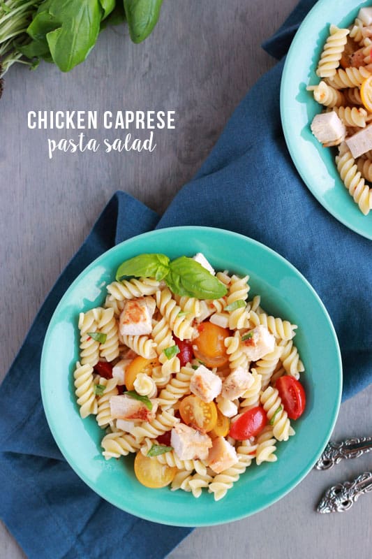 blue bowl filled with spiral pasta, grilled chicken pieces, sliced tomatoes and fresh basil on a dark blue napkin all sitting on a grey background