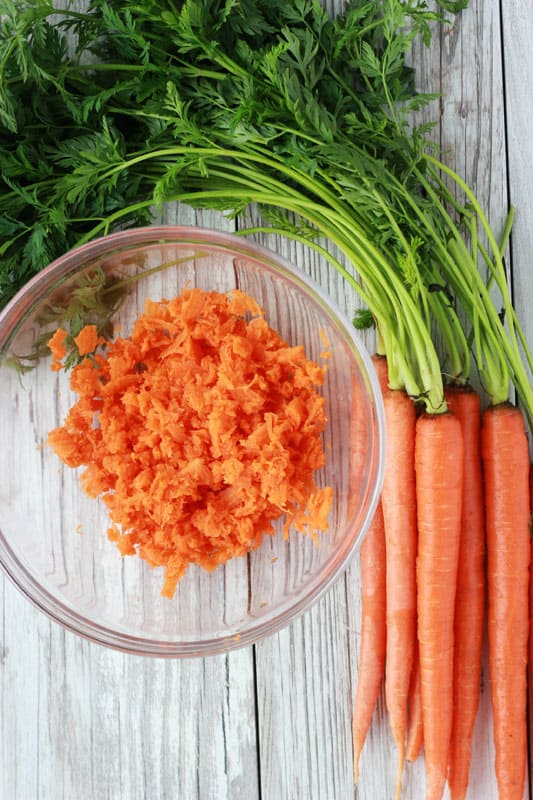 carrots shredded in a glass bowl with whole carrots on the table to the side