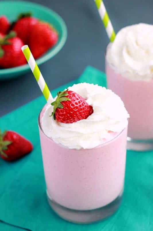 pink milkshake that is topped with whipped cream and a fresh strawberry with a green and white striped straw. The glass is sitting on a teal napkin with a bowl of strawberries off in the background