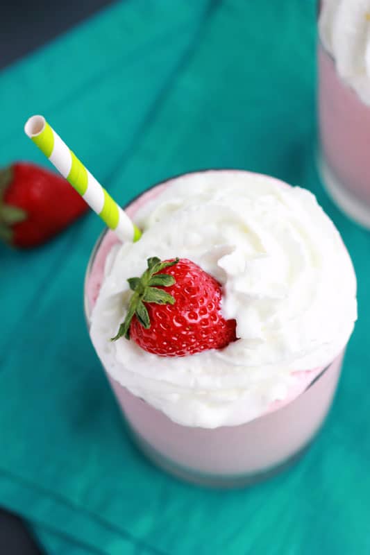 pink milkshake that is topped with whipped cream and a fresh strawberry with a green and white striped straw. The glass is sitting on a teal napkin with a bowl of strawberries off in the background