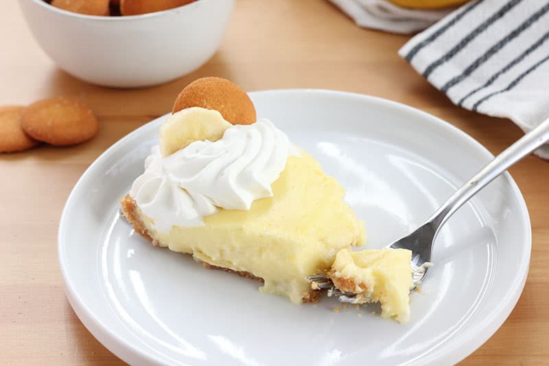 slice of pie on a white plate with a bite sitting on a fork. Topped with whipped cream swirl, vanilla cookie and slice of banana