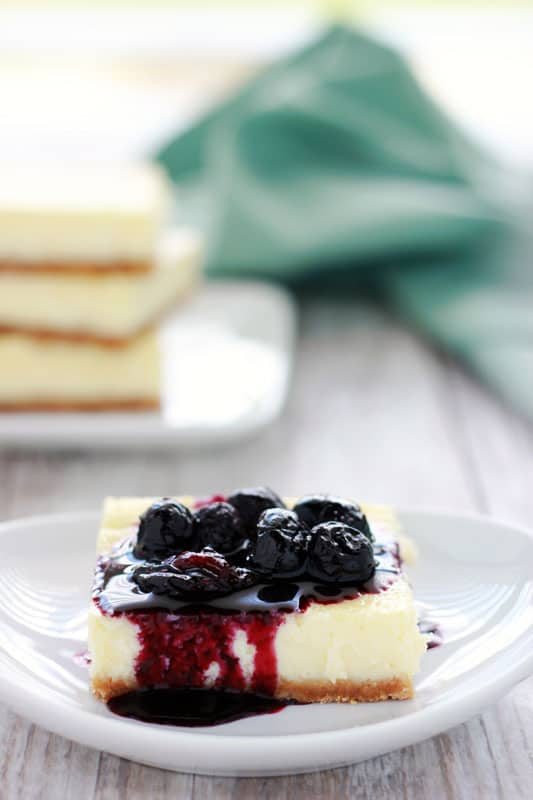 single cheesecake bar sitting on a white plate topped with a blueberry compote. Small stack of bars off in the background with a light blue napkin