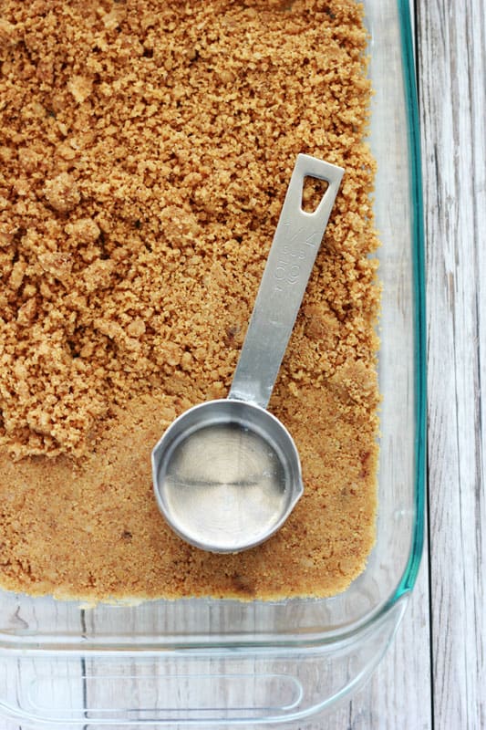 top down image showing a baking pan filled with graham crackers being pressed down with a metal measuring spoon