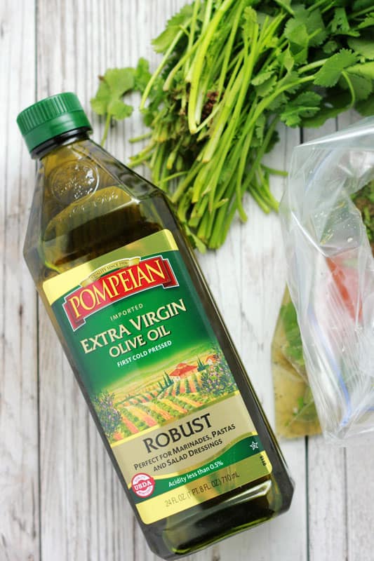 Pompeian Robust olive oil sitting on a white wooden table with fresh cilantro and a zip top bag