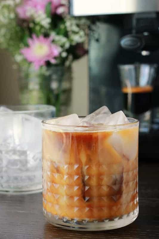 close up showing a short glass that is filled with an espresso shot and a swirl of milk on a dark table top. There is another short glass slightly behind the first that is filled with ice